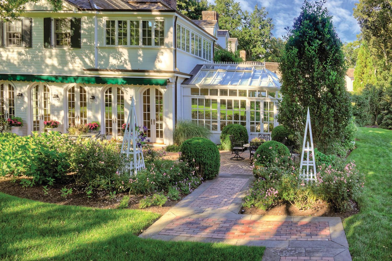 Brick walkway leading to a traditional residence with formal garden beds, shrubs, and lawn