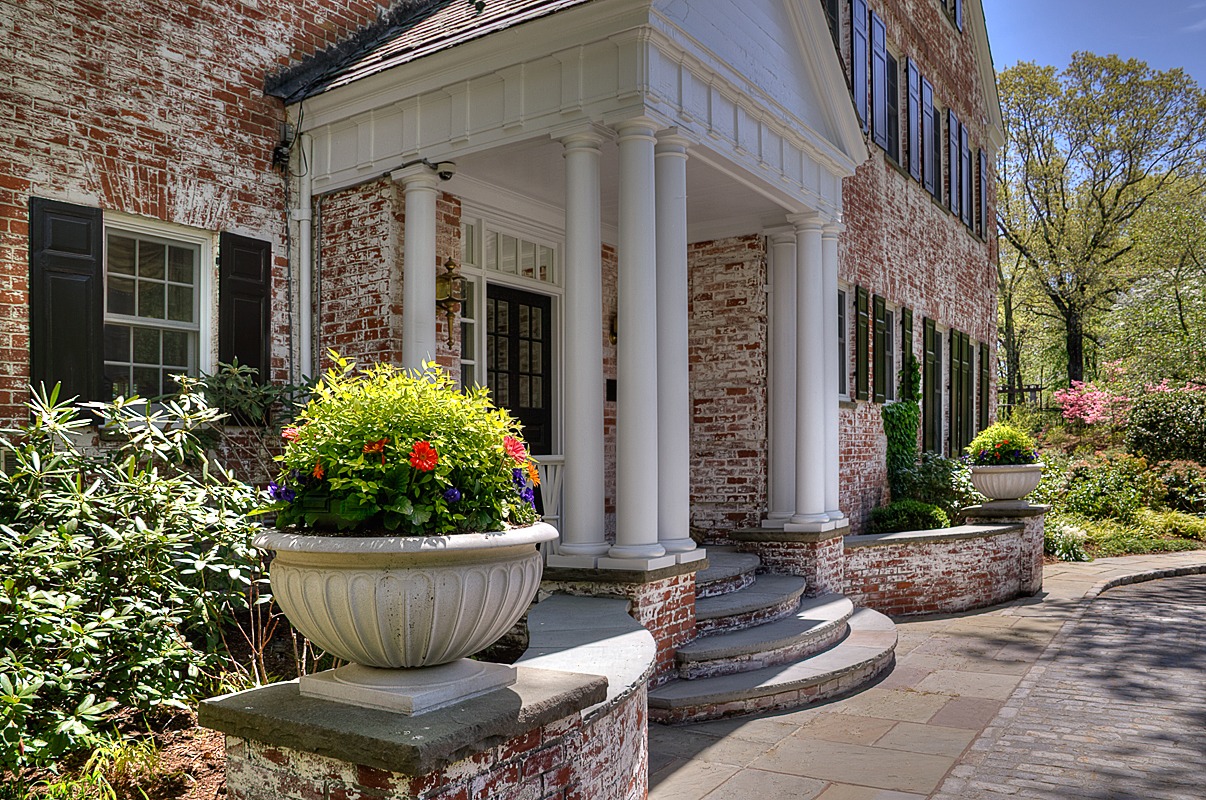 Charming brick colonial-style home with a weathered exterior, elegant white columned portico, and large decorative planters overflowing with colorful flowers, set along a curved stone walkway surrounded by lush greenery.