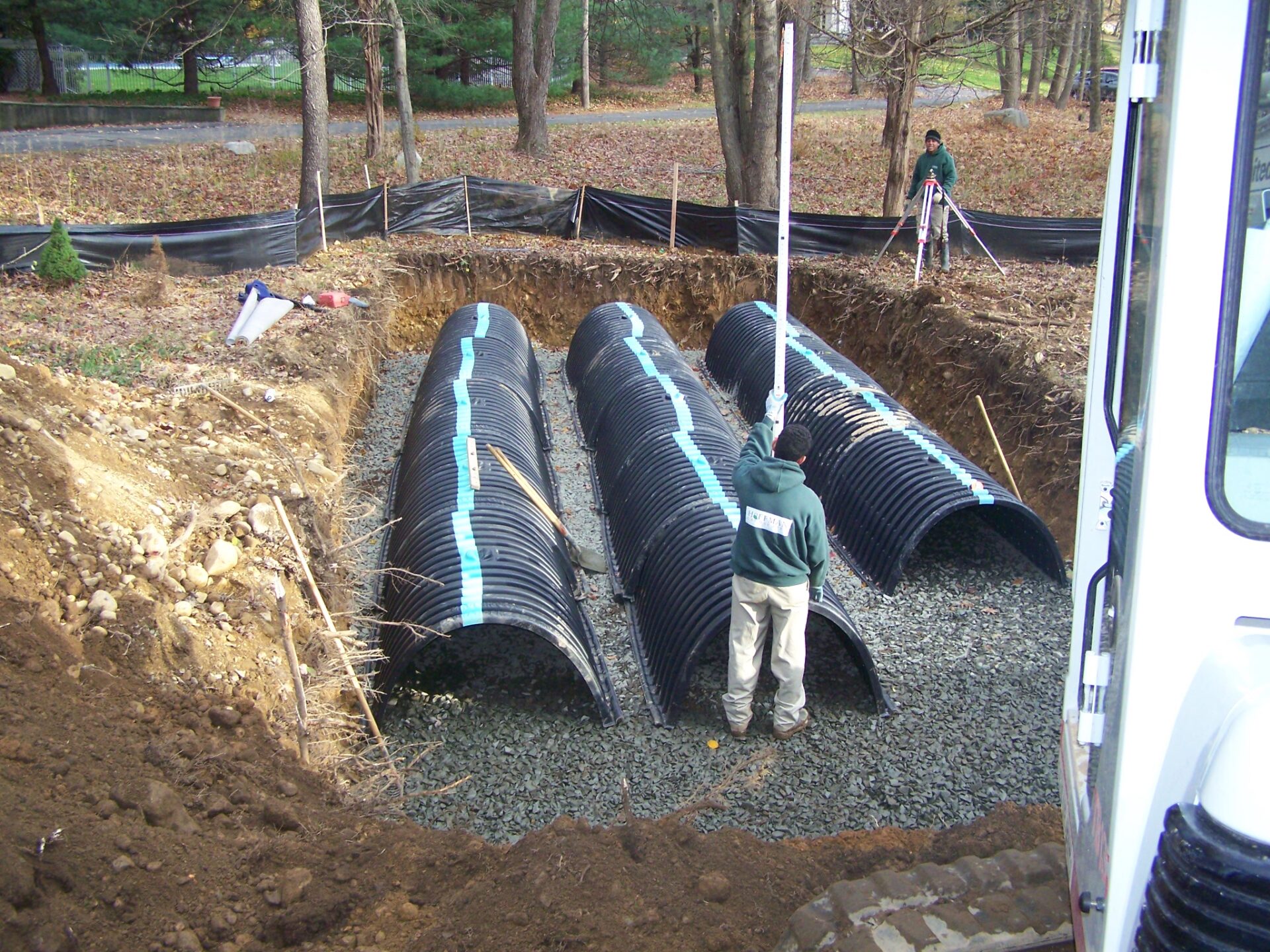 Two workers install a subsurface stormwater management system using black plastic infiltration chambers in a gravel trench surrounded by soil, with survey equipment and tools visible nearby.