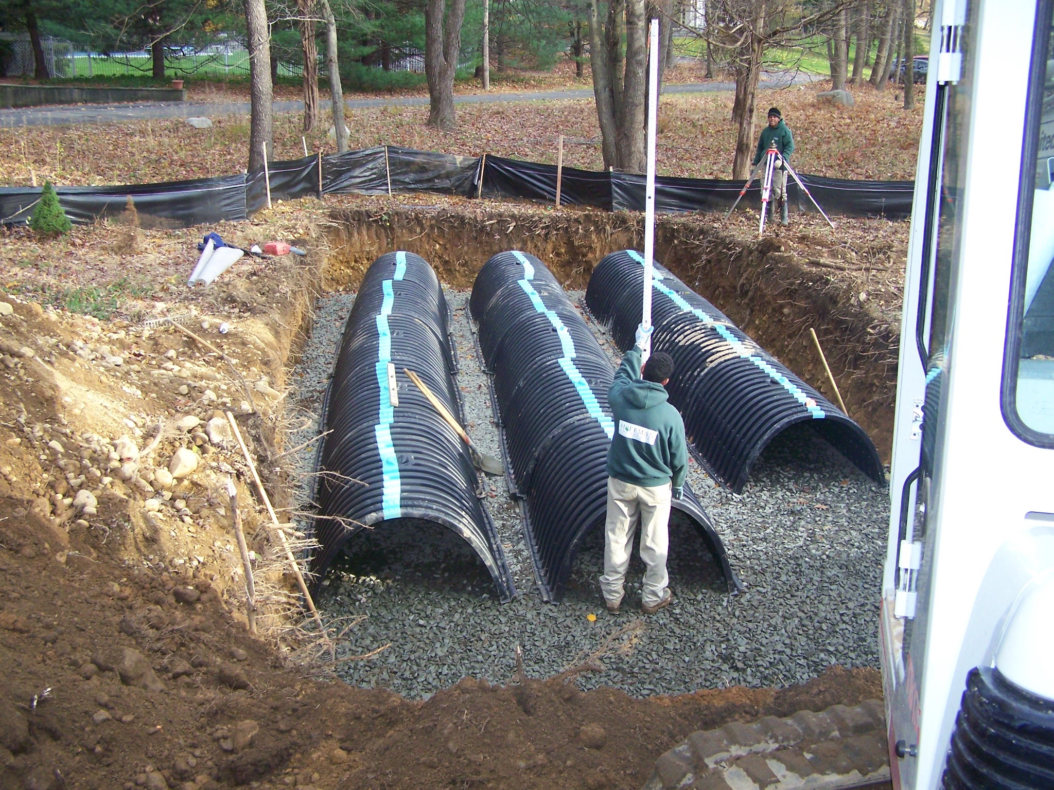 Two workers install a subsurface stormwater management system using black plastic infiltration chambers in a gravel trench surrounded by soil, with survey equipment and tools visible nearby.