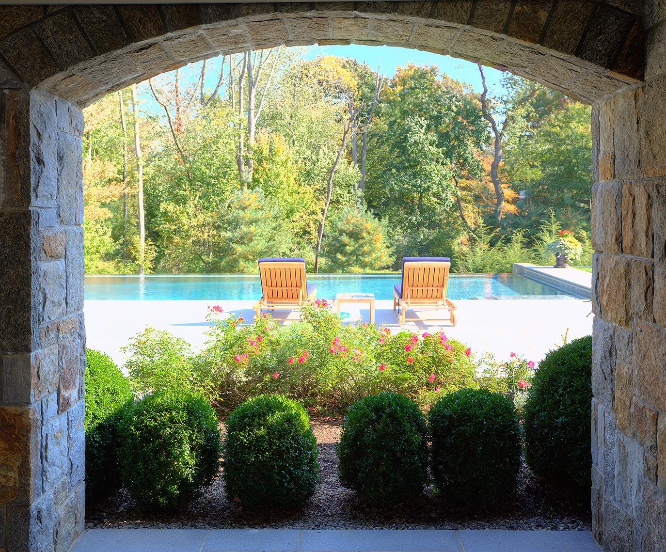 Stone archway framing a pool terrace with structured evergreen plantings and surrounding woodland